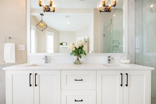 Bright and modern bathroom featuring a double vanity with white cabinets and black handles. A large mirror above the sinks reflects the space. There are two decorative light fixtures, a glass shower, and a vase with white flowers on the counter, courtesy of top Bathroom Remodelers Long Island.